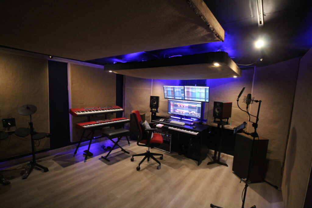 Professional recording studio with computer mixing area, dual monitors, Nord keyboards, and audio equipment.